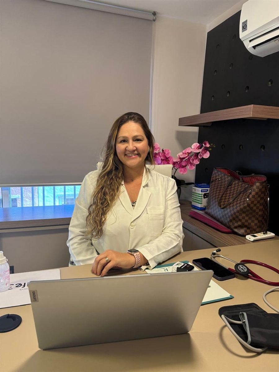 Dra. Raphaela Mannarino no consultório, de jaleco, diante do notebook e do estetoscópio