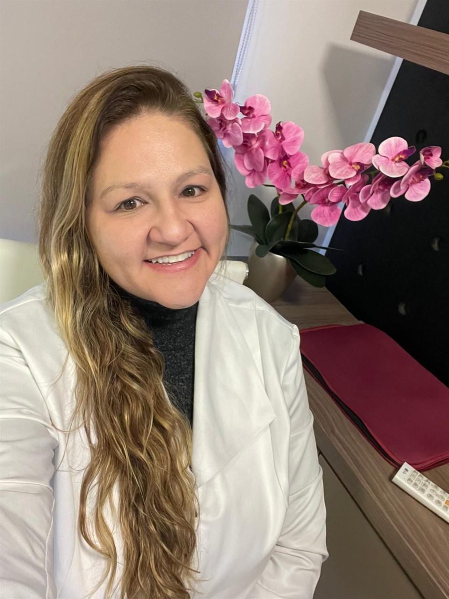 Dra. Raphaela Mannarino, cardiologista, de jaleco branco ao lado de um arranjo de orquídeas