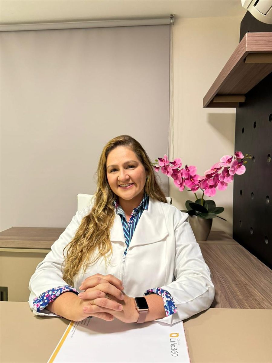 Dra. Raphaela Mannarino, cardiologista, de jaleco e mãos cruzadas sobre a mesa do consultório